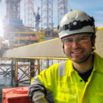 Een glimlachende offshore-medewerker in een felgele veiligheidsjas, uitgerust met een helm, hoofdlamp en veiligheidsbril. Op de achtergrond is een indrukwekkend boorplatform op open zee zichtbaar.